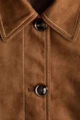 Chaqueta corta marrón de acabado cepillado suave con costuras en contraste. Cuenta con cuello clásico, cierre frontal de botones, bolsillos de parche delanteros y mangas largas.