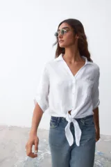 Camisa blanca de lino con cuello clásico, mangas 3/4 con botón para enrollar, bolsillo en el pecho y cierre frontal con botones. Se ata en la cintura.
