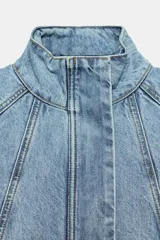Cazadora bomber de denim con cuello subido, manga larga y cierre frontal con cremallera y botones a presión ocultos. Presenta costuras verticales decorativas y bajo con ajuste elástico.