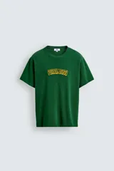 Camiseta de corte relajado, color verde, con cuello redondo y manga corta. Presenta un texto bordado en relieve con letras amarillas y contorno oscuro en el centro del pecho.