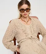 Blusa tipo bata de encaje texturizado color beige, con escote en V, mangas largas y volados aplicados en el escote. Presenta cierre frontal con botones y lazo de ajuste en la cintura.