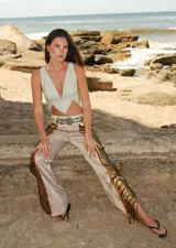 Pantalón de tiro medio con corte acampanado y tajos laterales. El tejido principal es brocato en tonos claros con un patrón sutil, y presenta apliques de lentejuelas doradas a lo largo de los tajos laterales. Incluye un cinturón ancho de estilo western con hebilla metálica.