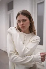Sweater de punto color crema, con cuello redondo y cierre frontal parcial con botones. Presenta mangas largas con puños acanalados y detalle de volados en los extremos.