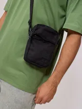 Bandolera negra de poliéster con logo de la marca Rusty en relieve en el frente. Cuenta con un bolsillo frontal con solapa y cierre de cremallera en el compartimento principal. La correa es ajustable.