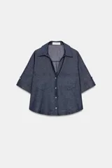 Camisa crop efecto denim color negro, con cuello solapa, escote en V, mangas cortas con trabilla y bolsillos frontales de parche. Cierre frontal con botones.
