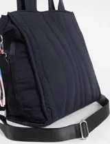 Cartera tipo shopper de color beige con diseño matelaseado de líneas onduladas y costuras en contraste azul. Cuenta con doble asa de hombro y un colgante decorativo de cordones multicolor en uno de sus laterales.