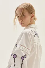 Blusa blanca de algodón con bordados geométricos en tonos negros y lilas, cuello clásico, cierre frontal con botones y mangas largas abullonadas con puños. Presenta detalles de vainillas y calados en el frente y mangas.