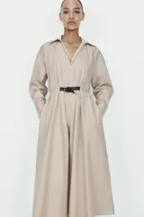 Vestido midi de color beige con cuello solapa y escote en pico. Presenta mangas largas, cintura elástica en la espalda y un cinturón a contraste con hebilla metálica. Incluye bolsillos laterales ocultos.