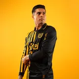 Camiseta de fútbol Puma de manga larga, conmemorativa del aniversario del Club Atlético Peñarol. Diseño de estilo retro que combina un sector con rayas verticales en amarillo y negro, y otro sector liso en negro, con cuello polo y detalles en dorado.