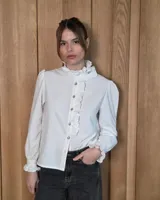 Camisa blanca de manga larga con cuello alto y volados decorativos en el cuello y la solapa frontal. Presenta cierre frontal con botones y puños con volados.