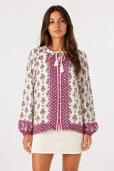 Blusa de viscosa con estampado floral y geométrico en tonos fucsia y verde sobre fondo crema. Presenta cuello redondo con cordones y borlas, cierre frontal con botones y mangas largas abullonadas con puños elásticos.
