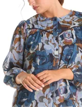 Blusa de gasa con estampado floral en tonos azules y marrones, con sutiles hilos de lurex dorados. Presenta cuello a la base, abertura central con botones, canesú delantero con pasamanería calada y mangas largas con puños elastizados. Incluye forro interno a tono.
