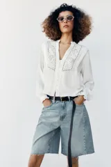 Blusa blanca de manga larga con cuello en V, cierre frontal con botones y detalles de bordados florales y encaje en el frente.