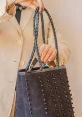 Cartera tipo tote de gamuza color marrón, con correas trenzadas y detalle de piedras naturales incrustadas en líneas verticales. Cuenta con cierre metálico y forro interior de tela.