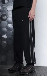 Pantalón deportivo de microfibra opaca, color negro, con diseño de vivos laterales blancos a contratono. Presenta cintura elástica y puños elásticos en los tobillos, con un pequeño logo bordado en la parte superior.