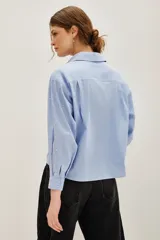 Camisa de corte recto y largo cropped, confeccionada en algodón con estampado de rayas verticales finas en color azul y blanco. Presenta cuello clásico, cierre frontal con botones y mangas largas con puños abotonados. La prenda está decorada con pequeñas aplicaciones de strass distribuidas por toda la superficie.