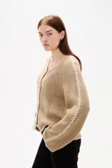 Cardigan de punto color beige con cuello en V, cierre frontal con botones y mangas largas holgadas. Presenta detalles de bordado decorativo en color blanco a lo largo de las mangas y los bordes.