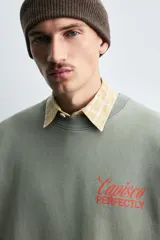 Sudadera de corte boxy fit, color verde oliva con efecto lavado. Presenta cuello redondo, manga corta terminada en rib y textos estampados a contraste en color rojo en el pecho.