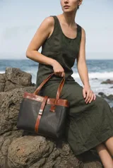 Cartera tipo tote de cuero vacuno, con diseño bicolor en gris y marrón. Cuenta con doble compartimento, cierre metálico, asas largas y una correa central decorativa con cierre de broche metálico. Incluye un detalle de etiqueta colgante de cuero.