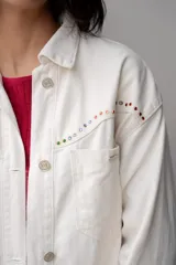 Chaqueta blanca de jean con cuello clásico, cierre frontal con botones metálicos y bolsillo de parche en el pecho. Presenta detalles de piedras de colores en el canesú.