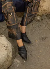 Botineta de gamuza azul marino con diseño drapeado, punta fina con puntera metálica y placa metálica decorativa en el lateral. Posee taco bajo cuadrado.