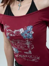 Remera bordó de manga corta con diseño de hombros descubiertos (off-shoulder) y un detalle superpuesto tipo bikini negro en el cuello. Cuenta con un estampado gráfico frontal de estilo vintage con flores y texto.