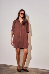 Vestido camisero corto de color marrón, con mangas cortas, cuello camisero, cierre frontal de botones y un bolsillo ojal en el pecho.
