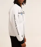 Campera tipo bomber blanca con detalles en negro, inspirada en uniformes espaciales de la NASA. Presenta parches con el logo de la NASA, la bandera de EE. UU. y texto en la manga que dice "Kennedy Space Center".