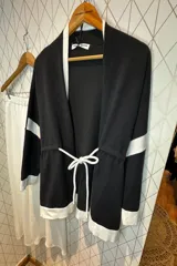 Cárdigan de punto en color gris con detalles en negro en los bordes, puños y una franja horizontal en las mangas. Presenta un diseño abierto con cordón ajustable en la cintura y bolsillos laterales.