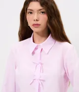 Camisa cropped de manga larga en tejido tricoline, con estampado de rayas verticales finas en color rosa y blanco. Presenta cuello camisero tradicional y cierre frontal decorado con tres lazos de tela. Diseño de corte corto y ajuste recto.