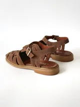 Sandalias estilo cangrejera color marrón, confeccionadas en cuero con diseño trenzado en la parte delantera, tira ajustable con hebilla metálica en el tobillo y suela plana.