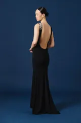 Vestido largo negro de corte sirena y silueta ajustada, con detalle de aplique floral brillante en el hombro izquierdo.
