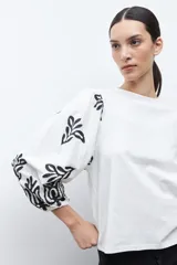 Remera de color crudo con mangas largas abullonadas, decoradas con bordados en color negro. Presenta un diseño de cuello redondo y corte recto.