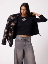 Campera reversible de diseño holgado, con un lado en tejido sherpa estampado con motivos florales en tonos negro y beige, y el otro lado en nylon negro. Cuenta con cierre frontal y cordones ajustables en el ruedo.