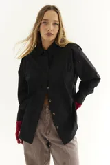 Camisa negra de popelina de algodón con cuello clásico, cierre frontal con botones y bolsillo en el pecho. Presenta detalles de zigzag en la parte delantera y trasera.