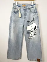 Jean de corte wide leg en denim azul claro con lavado desgastado. Presenta una ilustración pintada a mano de Snoopy en la pierna derecha, acompañada por la palabra 'Real', una corona y una estrella. Incluye etiqueta de cuero en la cintura y en el ruedo.