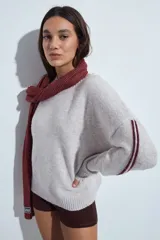 Sweater gris de calce relajado con cuello redondo y mangas largas. Presenta detalles de rayas horizontales en color rojo a lo largo de las mangas, con puños y ruedo acanalados.