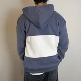 Canguro con capucha y cierre frontal parcial, de diseño color block en azul grisáceo y blanco. Presenta bordado con logo y texto en el pecho.