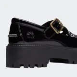 Zapato tipo Mary Jane de charol negro con plataforma gruesa y suela track. Presenta un diseño de tira con hebilla dorada y costuras blancas decorativas en la puntera. Incluye el logo de Timberland grabado en el talón.