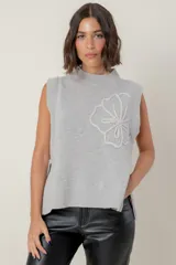 Chaleco de punto color gris, con cuello redondo, diseño sin mangas y detalle de flor bordada en el frente. Presenta tajos laterales en el ruedo.