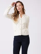 Cardigan blanco de punto aran con cuello redondo y cierre frontal de botones dorados. Presenta bolsillos simulados en la parte delantera.