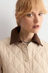 Campera capitoneada de diseño acolchado con patrón de rombos, color beige claro. Presenta cuello camisero de pana en color marrón oscuro, cierre frontal con botones metálicos y bolsillos frontales de parche.