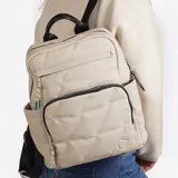 Mochila color beige con diseño acolchado en forma de rombos. Tiene un bolsillo frontal con cierre, un bolsillo lateral abierto y un compartimento principal con cierre. Las correas son ajustables y tiene un asa superior.