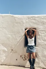 Top blanco estilo corsé con escote corazón, tirantes finos y bordados a tono. Presenta un bajo con volantes y cierre frontal con ganchos metálicos. La espalda es de tejido elástico tipo nido de abeja.