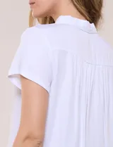 Camisa blanca de rayón con cuello camisero, escote en V y mangas cortas. Presenta abotonadura frontal con botones nacarados y detalles de tablas decorativas en el ruedo.