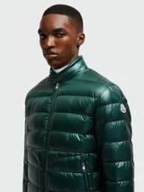 Campera corta tipo puffer de Moncler, modelo Acorus, en color verde oscuro con acabado brillante. Presenta diseño acolchado horizontal, cuello alto con cierre de broche, cierre frontal completo y bolsillos laterales con cierre.