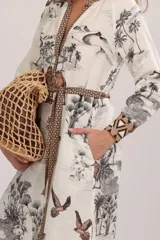 Túnica midi color blanco de manga larga, con escote en V profundo y lazo ajustable en la cintura. Presenta un estampado de paisaje desértico con palmeras y camellos, además de detalles geométricos en el escote, puños y cinturón.