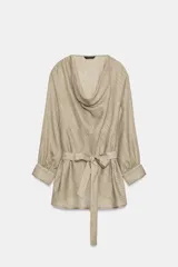 Camisa de cuello fluido y manga larga con puños anchos, de corte recto y largo medio, confeccionada en tejido rústico en tono beige con sutil textura jaspeada. Cuenta con un cinturón ajustable del mismo género para entallar la cintura.