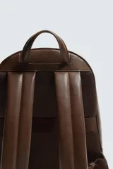 Mochila lisa de color marrón oscuro con acabado mate. Cuenta con un compartimento principal con cierre de cremallera, un bolsillo interior con cremallera y un bolsillo frontal exterior también con cremallera. La parte posterior es acolchada e incluye una banda de sujeción para equipaje. Dispone de un asa de mano superior y dos asas de hombro ajustables.