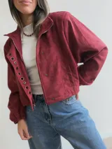 Chaqueta corta de efecto gamuza en color azul marino, con cierre frontal y solapa decorada con una hilera de aros metálicos. Presenta mangas tipo globo, ajuste lateral en la cintura y forro interior.